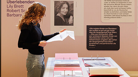 Frau schaut sich in einer Ausstellung Dokumente an, die in riesigen großen roten Ringordnern abgeheftet sind. Die Ordner liegen auf einem Tisch.
