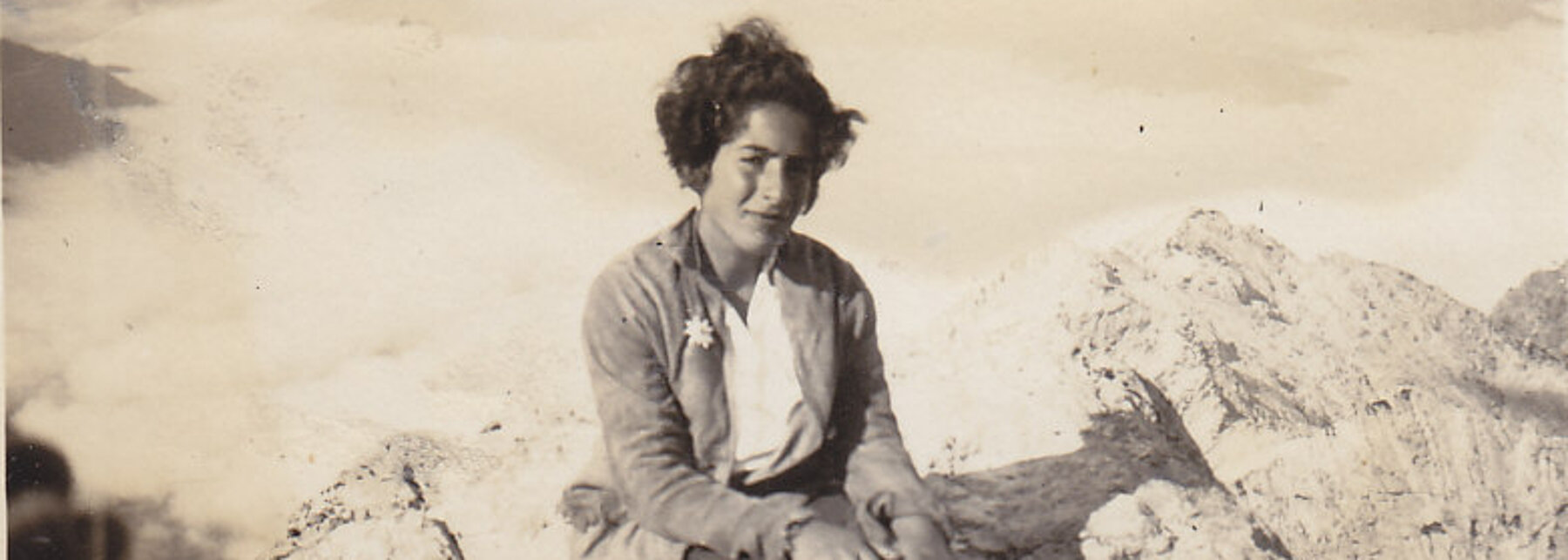 Schwarz-Weiß-Fotografie: Eine junge Frau, Grete Weil, sitzt hoch oben auf einem Gipfel. Ihre kurzen dunklen Haare flattern im Wind. Sie trägt bayrische Wanderkleidung mit Edelweiß im Knopfloch.