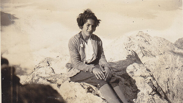 Schwarz-Weiß-Fotografie: Eine junge Frau, Grete Weil, sitzt hoch oben auf einem Gipfel. Ihre kurzen dunklen Haare flattern im Wind. Sie trägt bayrische Wanderkleidung mit Edelweiß im Knopfloch.