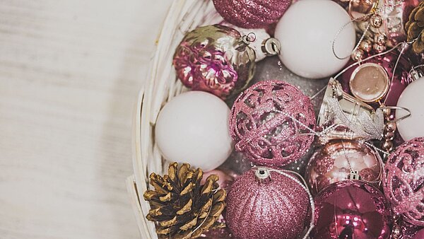 Weißer Korb auf hellem Holz mit rosa-weißen Weihnachtskugeln und einem Tannenzapfen.