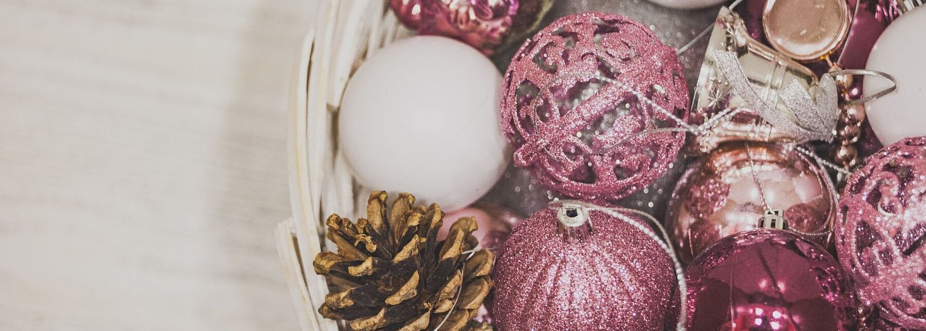 Weißer Korb auf hellem Holz mit rosa-weißen Weihnachtskugeln und einem Tannenzapfen.