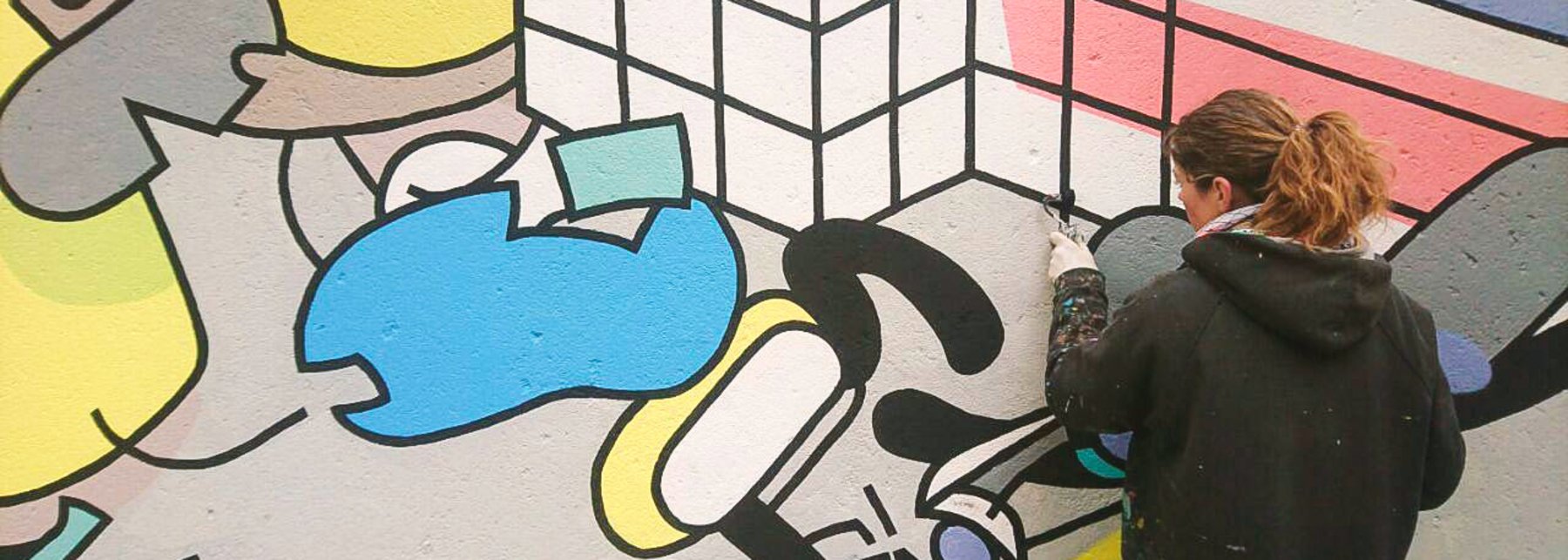 Eine junge Frau steht an einer bereits bunt bemalten Wand und gestaltet dort das Bild weiter