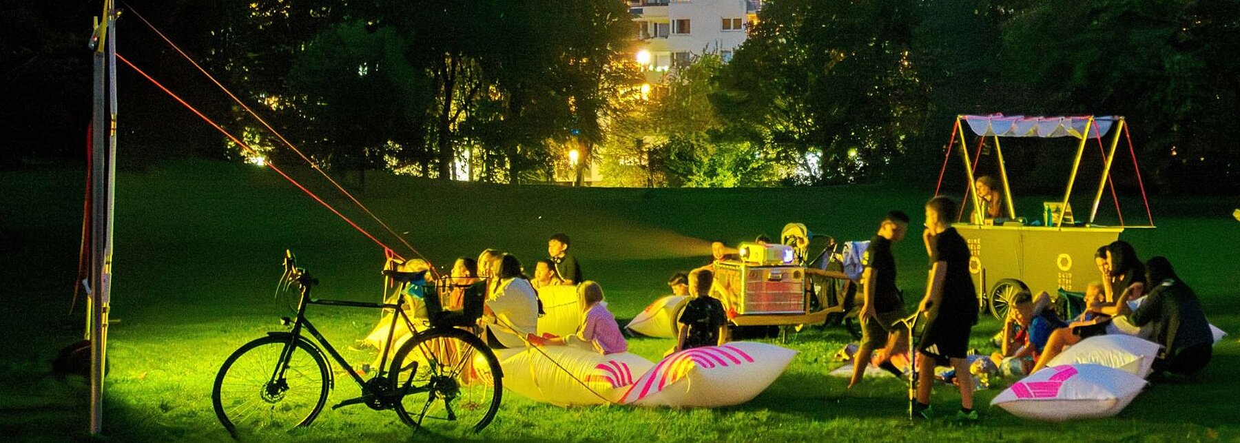 Aufgespannte Leinwand auf einer Wiese, abendliche Stimmung, der Beamer auf dem Lastenfahrrad projeziert einen Film auf die Leinwand. Menschen sitzen auf der Wiese und schauen zu