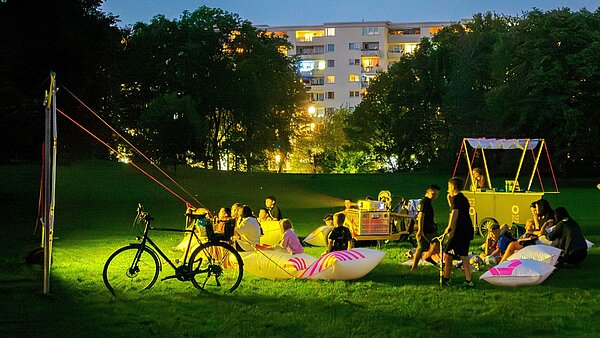 Aufgespannte Leinwand auf einer Wiese, abendliche Stimmung, der Beamer auf dem Lastenfahrrad projeziert einen Film auf die Leinwand. Menschen sitzen auf der Wiese und schauen zu