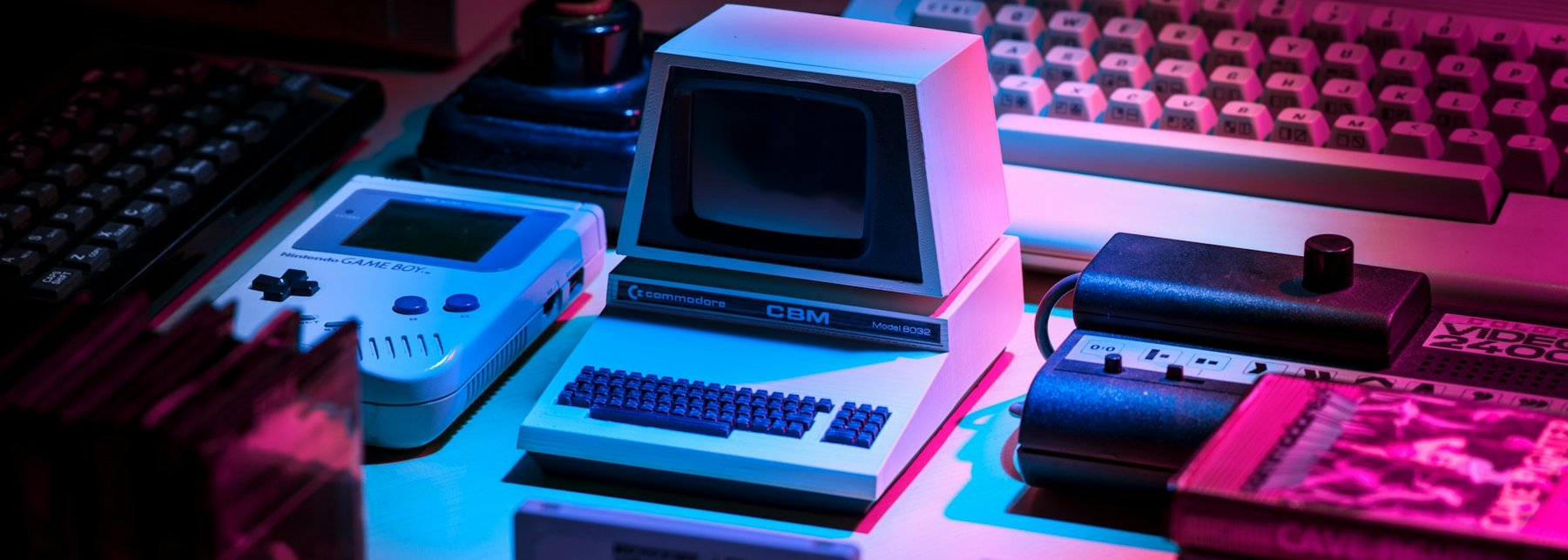 Mehrere überholte Konsolen, Datenträger und Computer geordnet auf einem Tisch mit pinken Licht beschienen.